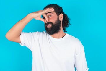 Displeased young bearded man wearing white T-shirt over blue studio background plugs nose as smells...