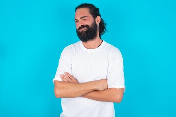 Picture of angry young bearded man wearing white T-shirt over blue studio background crossing arms. Looking at camera with disappointed expression.