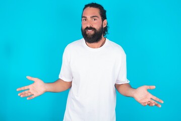 young bearded man wearing white T-shirt over blue studio background looks uncertain shrugs...