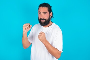 Portrait of attractive young bearded man wearing white T-shirt over blue studio background holding hands in front of him in boxing position going to fight.