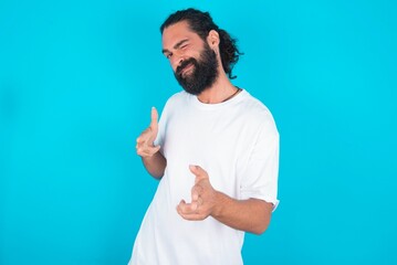 Hey you, bang. Joyful and charismatic good-looking young bearded man wearing white T-shirt over blue studio background winking and pointing with finger pistols at camera happily and cheeky. - obrazy, fototapety, plakaty