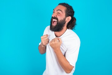 young bearded man wearing white T-shirt over blue studio background excited and glad to achieve victory, clenches fists, screams in excitement with closed eyes,successful person.