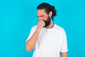young bearded man wearing white T-shirt over blue studio background, holding his nose because of a bad smell.