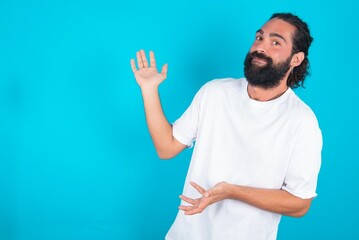 young bearded man wearing white T-shirt over blue studio background pointing aside with both hands showing something strange and saying: I don't know what is this. Advertisement concept.