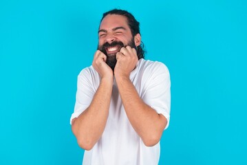 Portrait of young bearded man wearing white T-shirt over blue studio background being overwhelmed, expressing excitement and happiness with closed eyes and hands near face.