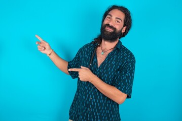 young bearded man wearing blue shirt over blue studio background with positive expression, indicates with fore finger at blank copy space for your promotional text or advertisement.