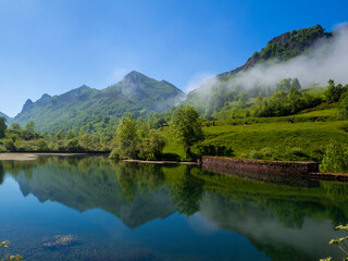 Fototapeta premium Vistas panorámicas de un paisaje con un lago, reflejos de las montañas y el cielo azul, con niebla blanca y árboles verdes en Asturias, España, verano de 2021.