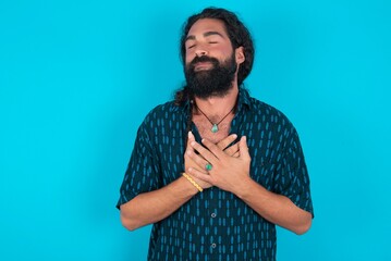 young bearded man wearing blue shirt over blue stud closes eyes and keeps hands on chest near heart, expresses sincere emotions, being kind hearted and honest. Body language and real feelings concept.