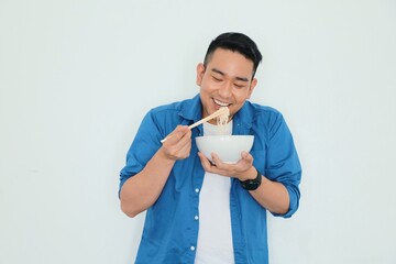 Happy Asian man in blue shirt holding Japanese noodle bowl and chopsticks standing over white background with copy space.Asian man eating Japanese noodle.