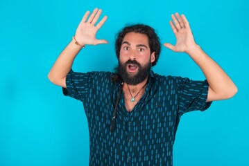 Caucasian man with beard wearing blue shirt over blue background looks with excitement at camera, keeps hands raised over head, notices something unexpected, reacts on sudden news.