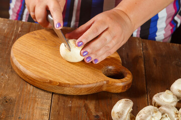 A woman cuts champignon mushrooms with a knife on a round wooden board.