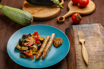 A plate of vegetables with grilled sausages on the table next to zucchini on wooden boards and a wooden fork.