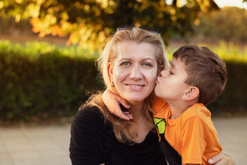 Fototapeta premium The little boy kisses his mother on the cheek, the mother is simply beaming with joy, both are happy and blessed to communicate. A woman and her son are resting and talking in the park