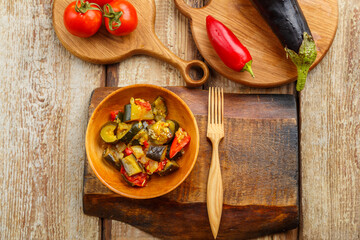 A plate of grilled vegetables next to tomatoes and eggplant and peppers on wooden boards next to a wooden fork.