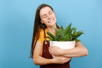 Dreamful young female closes eyes embraces pot of houseplant, new flower for home garden, posing...