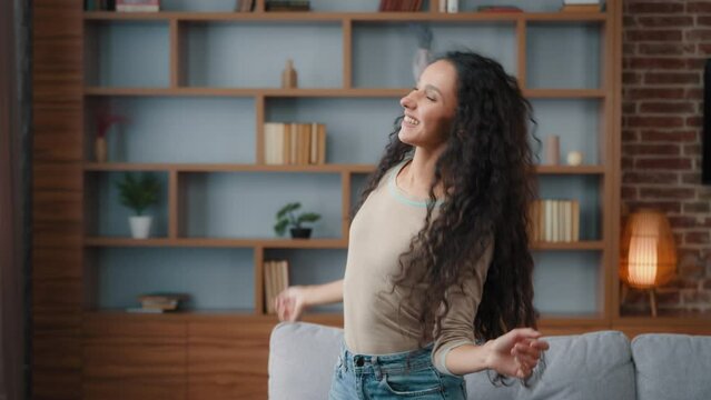 Graceful Caucasian girl dancer at home in living room dancing to music Hispanic curly haired woman enjoying weekend moving to rhythm of favorite musical song funny dance having fun alone move hands