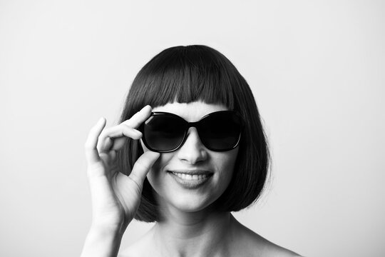 Beauty, fashion concept. Portrait of beautiful and sexy woman with bob style wig and sunglasses looking to camera with smile. Model with naked shoulders and white teeth. Black and white image
