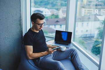 A young businessman is looking for something and typing on a smartphone and drinking fresh black coffee. The concept of a modern successful person. Young focused guy with glasses in an open office