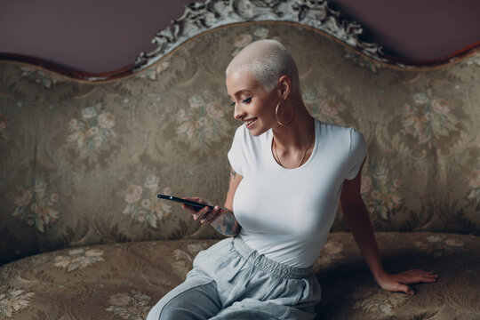 Millenial young woman with short blonde hair portrait sitting and texting message with mobile phone on vintage sofa