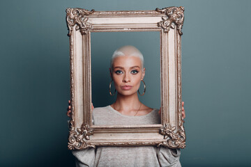 Millenial young woman with short blonde hair holds gilded picture frame in hands behind her face...
