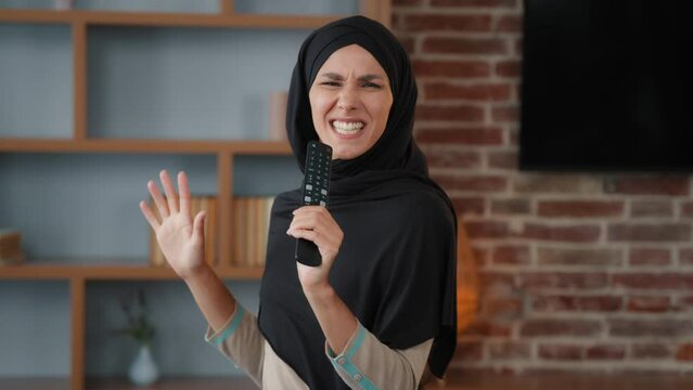 Arabian Muslim Islamic woman happy funny girl in hijab at home singing song in TV remote controller like microphone sing cheerful favorite sound vocal karaoke indoor in living room dancing having fun
