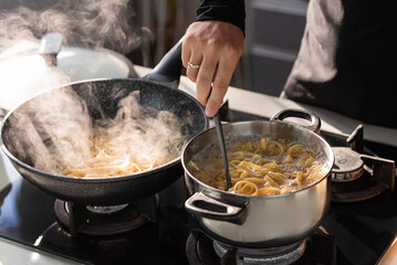 Professional chef cook making Italian Tagliatelle pasta with mushrooms and cream at modern kitchen gas stove in wok pan and pot.