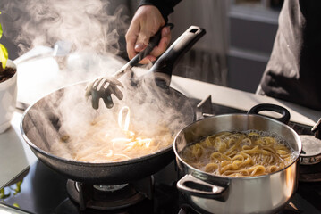 Professional chef cook making Italian Tagliatelle pasta with mushrooms and cream at modern kitchen gas stove in wok pan and pot.