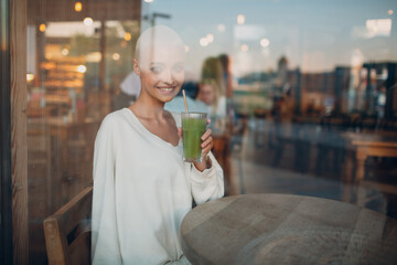 Portrait of young smiling millenial european short haired woman with green smoothie behind window glass at cafe. Beautiful happy blonde girl indoor.