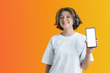 cheerful cute teenager girl with curly hair in trendy glasses holds smartphone in her hand with an empty white screen on colored background, for banner design, business cards and mockup