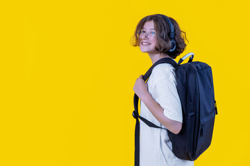 portrait of joyful caucasian teenage girl with glasses, headphones and backpack on her back on colored background, back to school, traveling with ease, studio photography