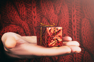 man in red sweater holds in his hand small red Christmas gift with bow, close-up, toned image