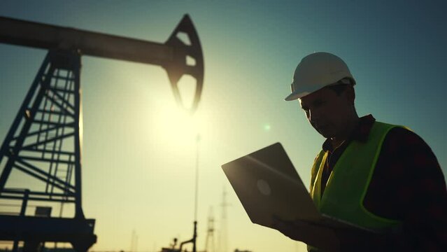 oil industry. a worker works next to an oil pump holding a laptop. industry business oil and gas concept. engineer studying the level of oil production on a laptop silhouette at sunset lifestyle