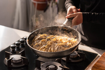 Professional chef cook making Italian Tagliatelle pasta with mushrooms and cream at kitchen gas stove in wok pan. Flying pasta levitation in motion.