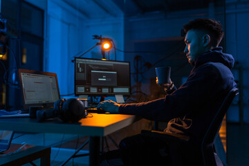 cybercrime, hacking and technology concept - male hacker in dark room drinking coffee and using...