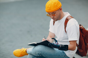 Young disabled man writes in notebook with pen in artificial prosthetic hand at city street outdoors