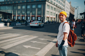 Young traveler disabled man with artificial prosthetic hand in casual clothes and backpack walking at sunny city street outdoor crosswalk arrows way