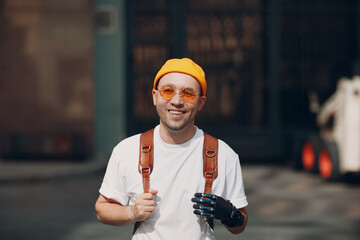 Young disabled man with artificial prosthetic hand in casual clothes and backpack walking at sunny city street outdoor