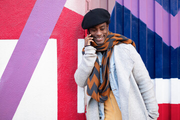 Young adult African American man having a friendly phone conversation. He is leaning against a colorful wall.