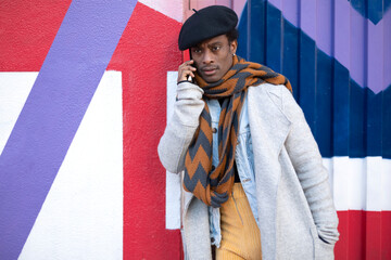 Young adult African American man talking on the phone with serious gesture. He is leaning against a colorful wall.