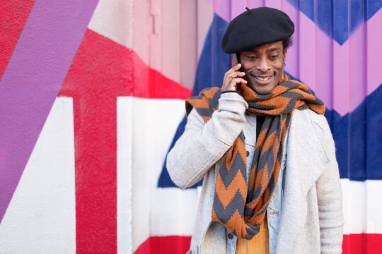 Young adult African American man talking on phone outdoors. He is standing next to a colorful wall. Space for text. - Powered by Adobe