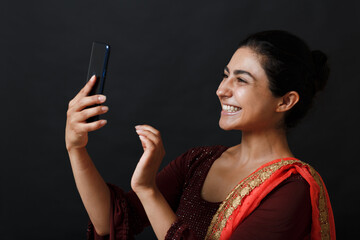 Happy indian woman having video call on mobile phone. Brunette in traditional sari clothes of red and gold colour. Beautiful smiling dark haired young woman. Isolated on dark background. Studio shoot