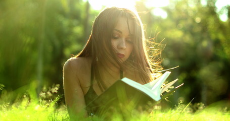 Woman reading book lying in grass at park. Pretty girl enjoying story novel in the sunlight
