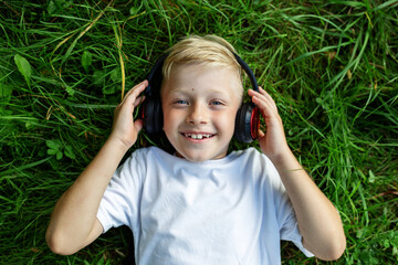 Child listens to music. Blond boy uses headphones and smart phone, lies in grass. Top view