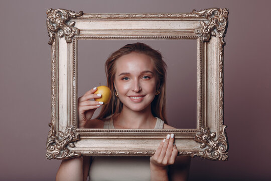 Millenial young woman blonde hair holds gilded picture frame in hands face portrait with apple