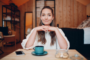 Smiling happy young woman sitting at table at home behind computer laptop and talking on video call. Girl female with cup of tea or coffee speaking online on webcam indoors