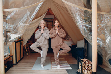 Fototapeta na wymiar Woman senior and young relaxing at glamping camping tent. Women family elderly grandmother and young granddaughter doing yoga and meditation indoor. Modern zen-like vacation lifestyle concept