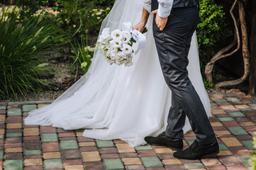 Plakat A stylish groom in a blue suit and a bride in a long dress are walking in the park outdoors with a bouquet of white roses in their hands. Wedding photography of the newlyweds, portrait.