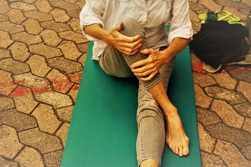 Auf grüner Gummimatte sitzende Frau in grauer Hose und weißer Bluse bei Yogaübung im Freien auf...