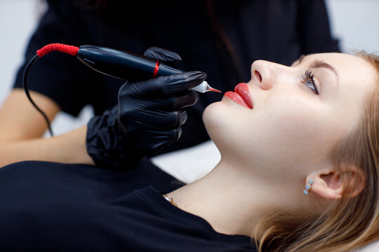 Cosmetologist applying pink pigment permanent tattoo on female lips with tatooing needle machine
