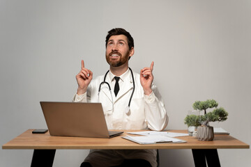 Handsome family doctor wearing white lab coat and stethoscope smiling friendly and pointing up